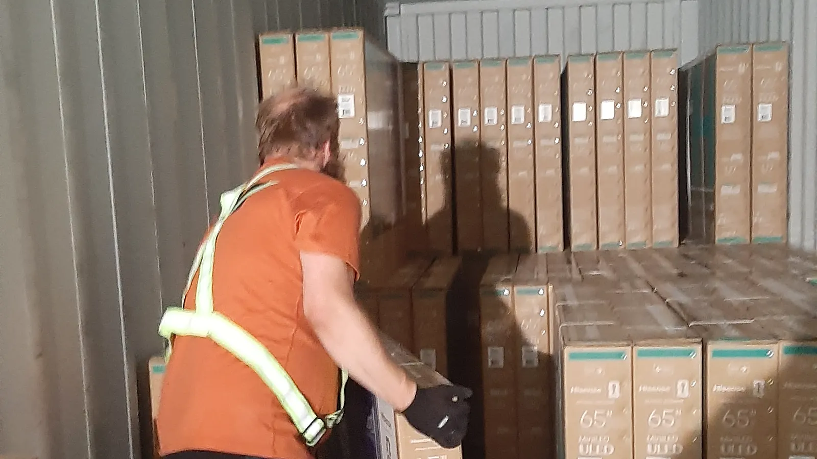 Crew member in a hi-vis vest hand-unloading large flat cartons inside a floor-loaded shipping container