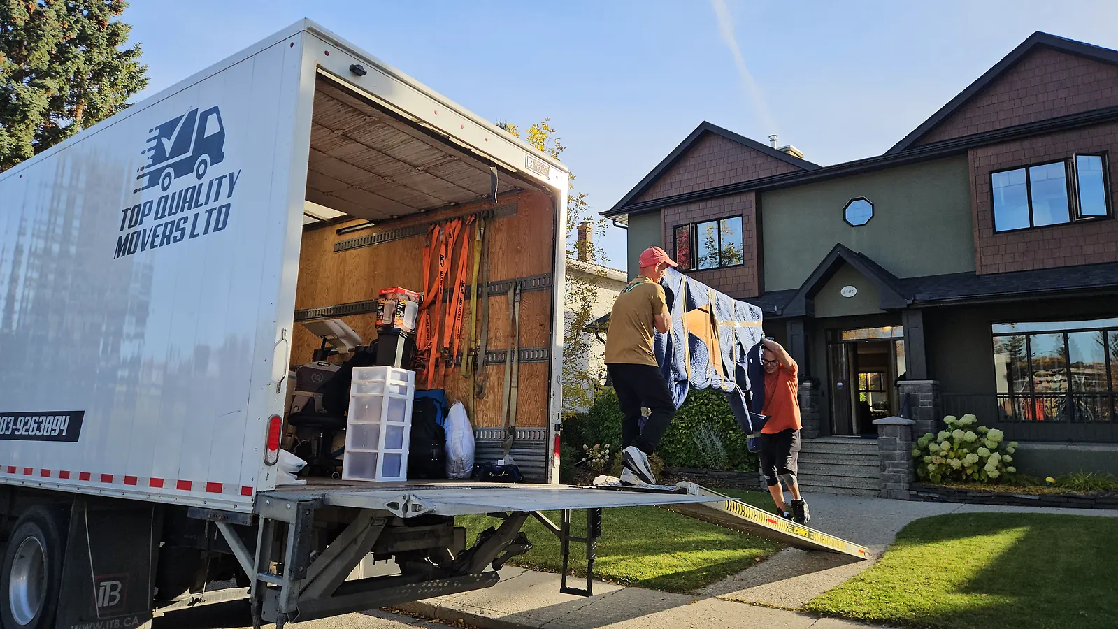 Two movers carrying a blanket-wrapped piece up the ramp into a Top Quality Movers truck at a two-storey house in Calgary
