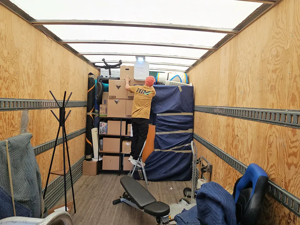 A mover on a step ladder stacking boxes high inside the truck, beside blanket-wrapped furniture and a weight bench