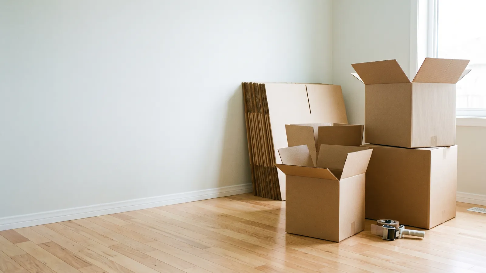 Moving boxes of different sizes, assembled with their flaps open, beside a bundle of flat-packed boxes and a tape gun in an empty room