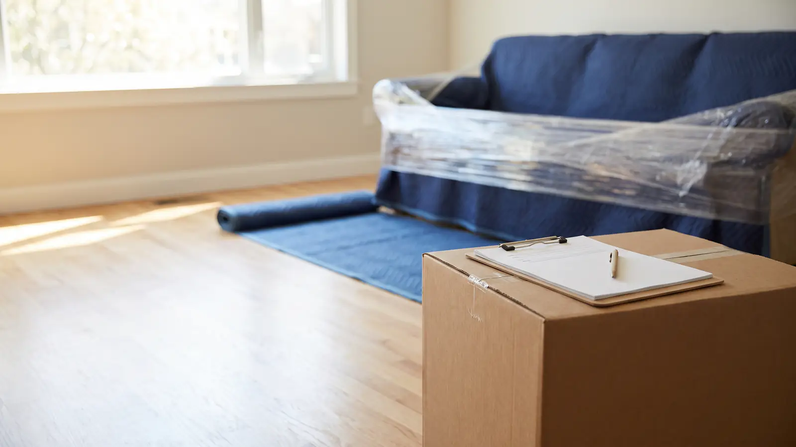 Moving insurance: a sofa wrapped in moving blankets and shrink wrap, with a clipboard on a sealed box in front
