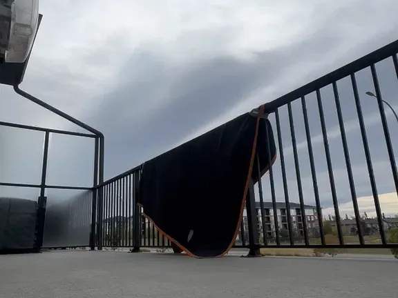 A black moving blanket with orange edging draped over a balcony railing, with apartment buildings in the distance