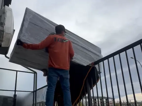 A crew member in an orange hoodie holding a shrink-wrapped couch up on the balcony railing