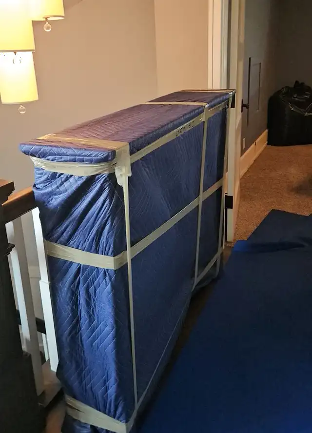 A tall piece of furniture wrapped in moving blankets and tape, standing on a floor runner at the top of the stairs in a home