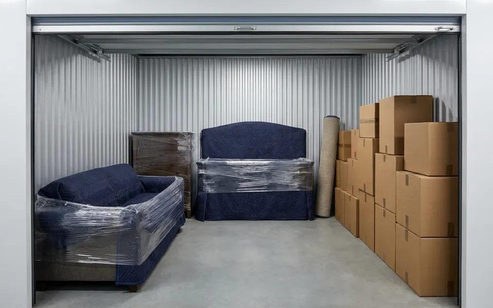 Furniture wrapped in moving blankets and shrink wrap beside stacked moving boxes in a storage unit