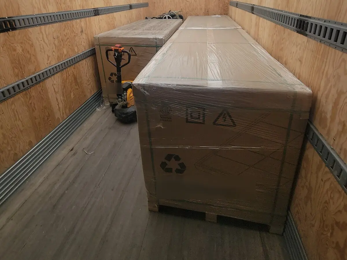 Freight delivery load: two long plastic-wrapped crates on pallets in a truck's cargo box, with an electric pallet jack