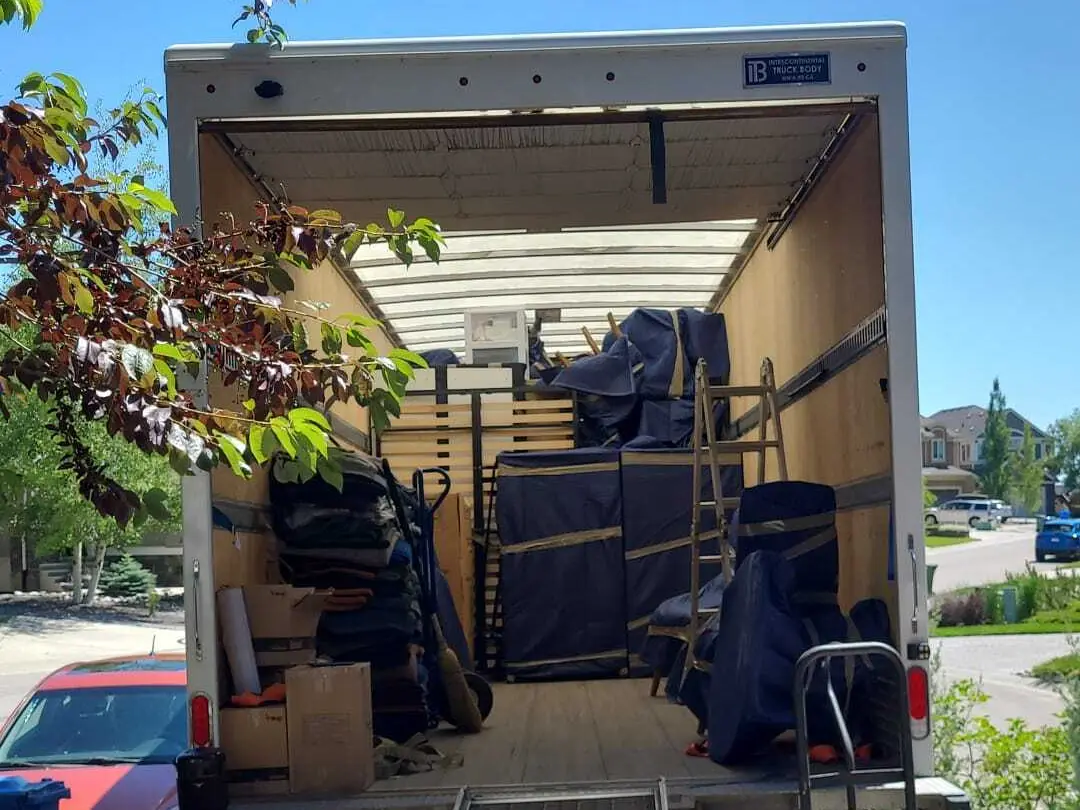 An open moving truck on a residential street, loaded with furniture wrapped in blue moving blankets